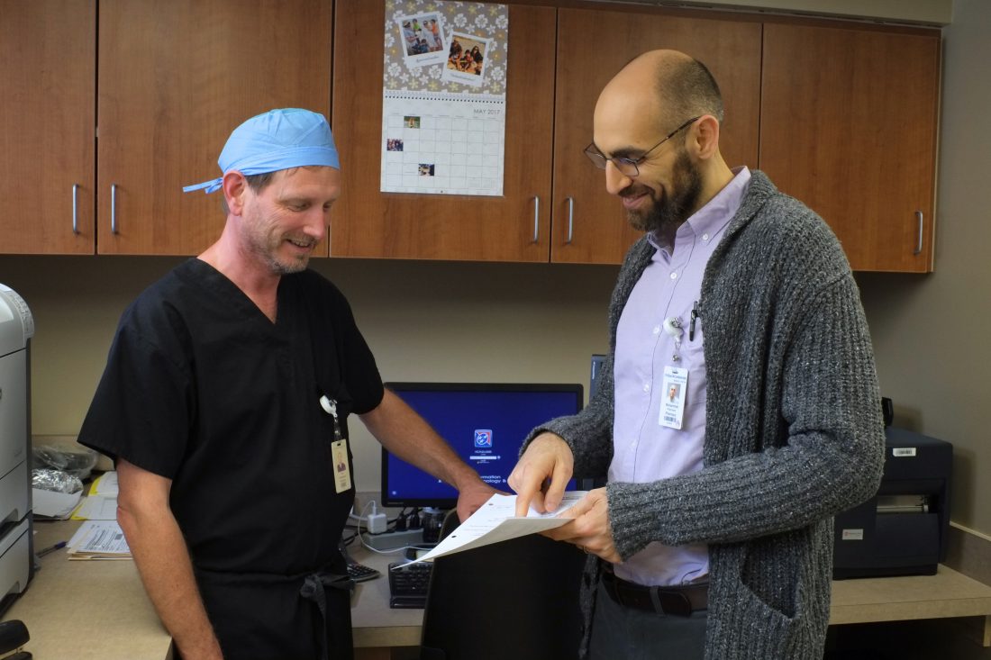 Man wearing button down shirt and sweater showing document to man wearing scrubs and a surgical cap