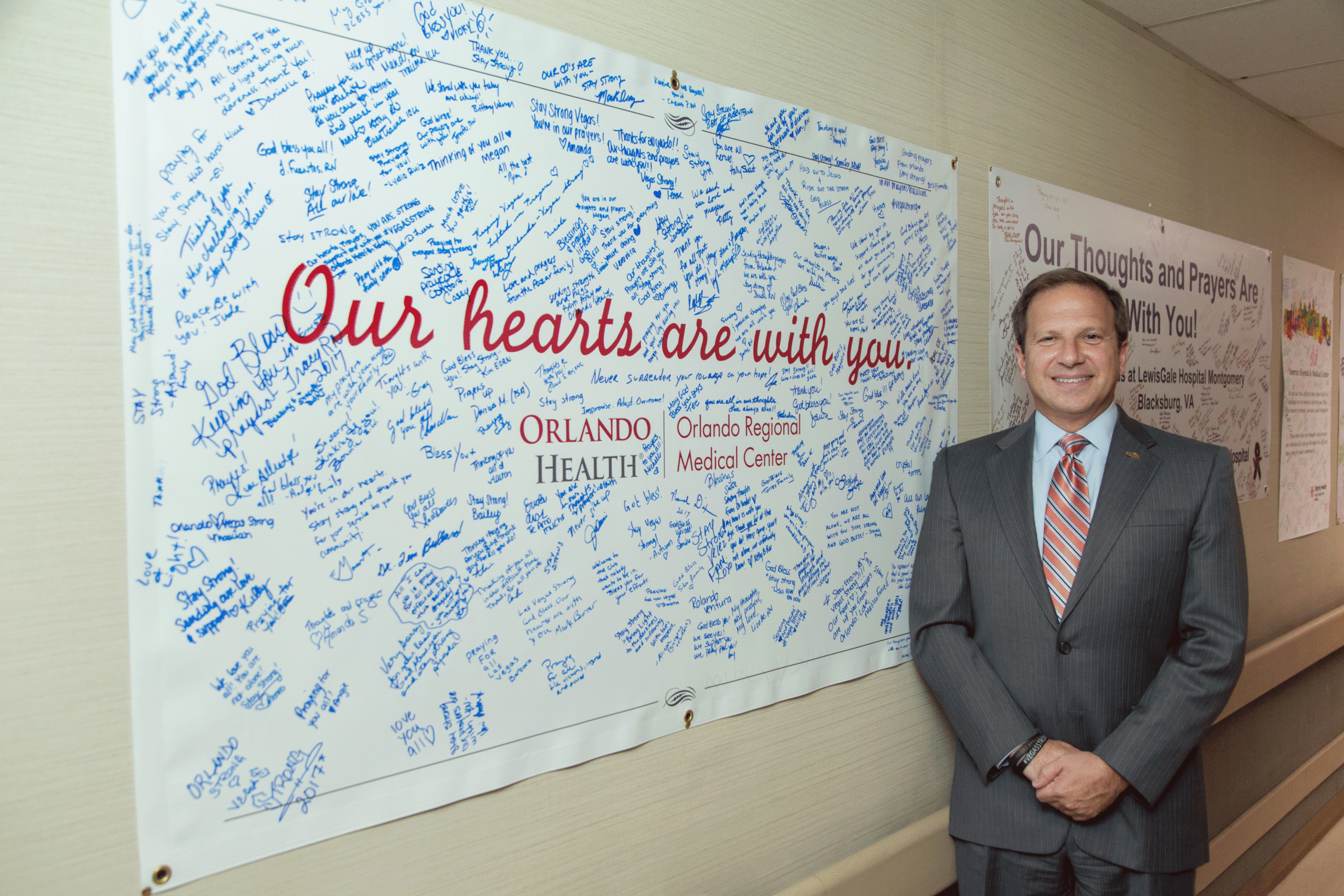 Man wearing suit and tie standing next to a poster filled with signatures that says Our Hearts Are With You and Orlando Regional Medical Center