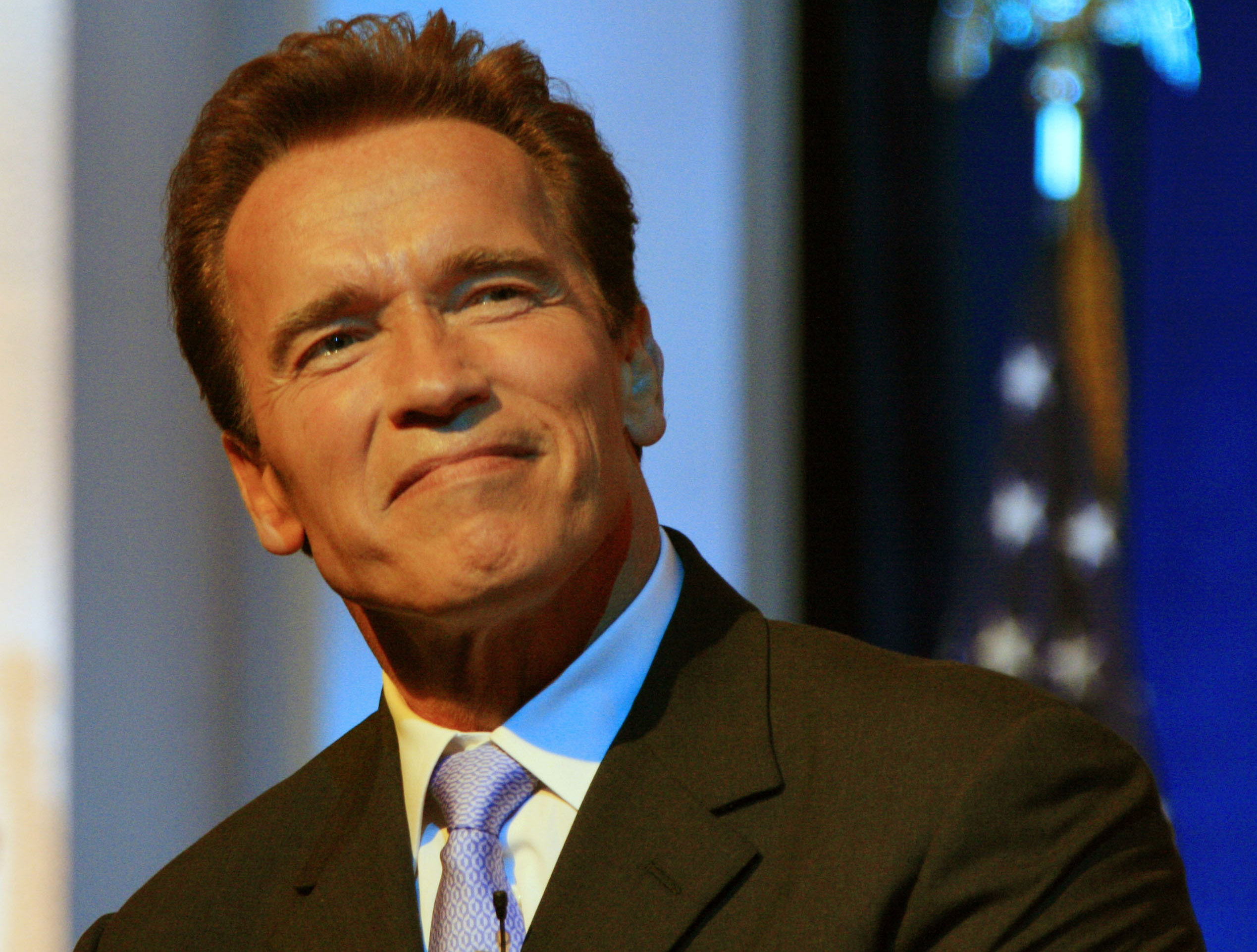 Arnold Schwarzenegger wearing suit and tie with American flag in the background