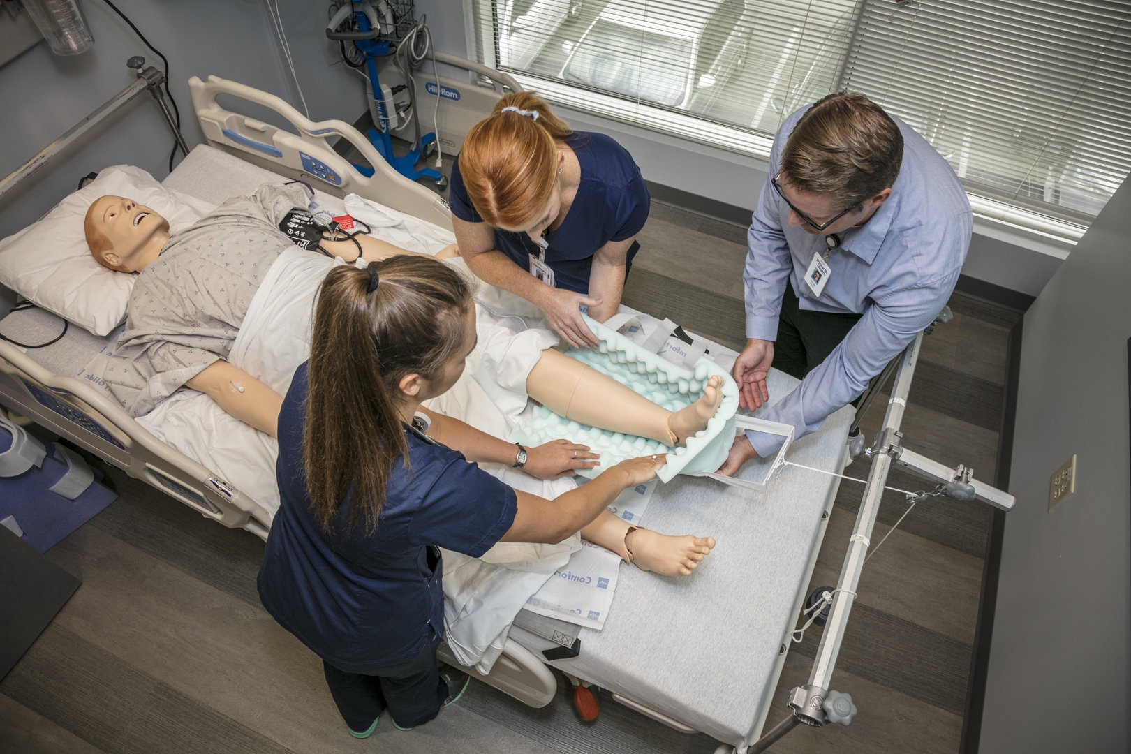 Two female nurses and a male instructor practice care on a mannequin in a hospital bed