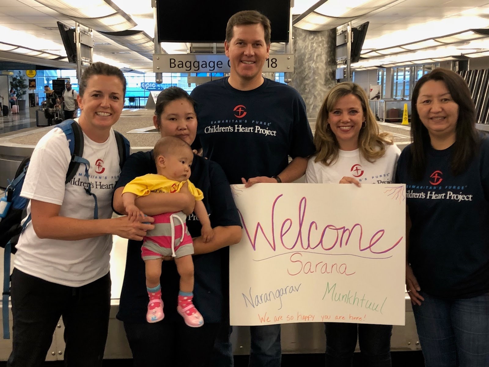 Five adults and a baby at an airport with a welcome sign