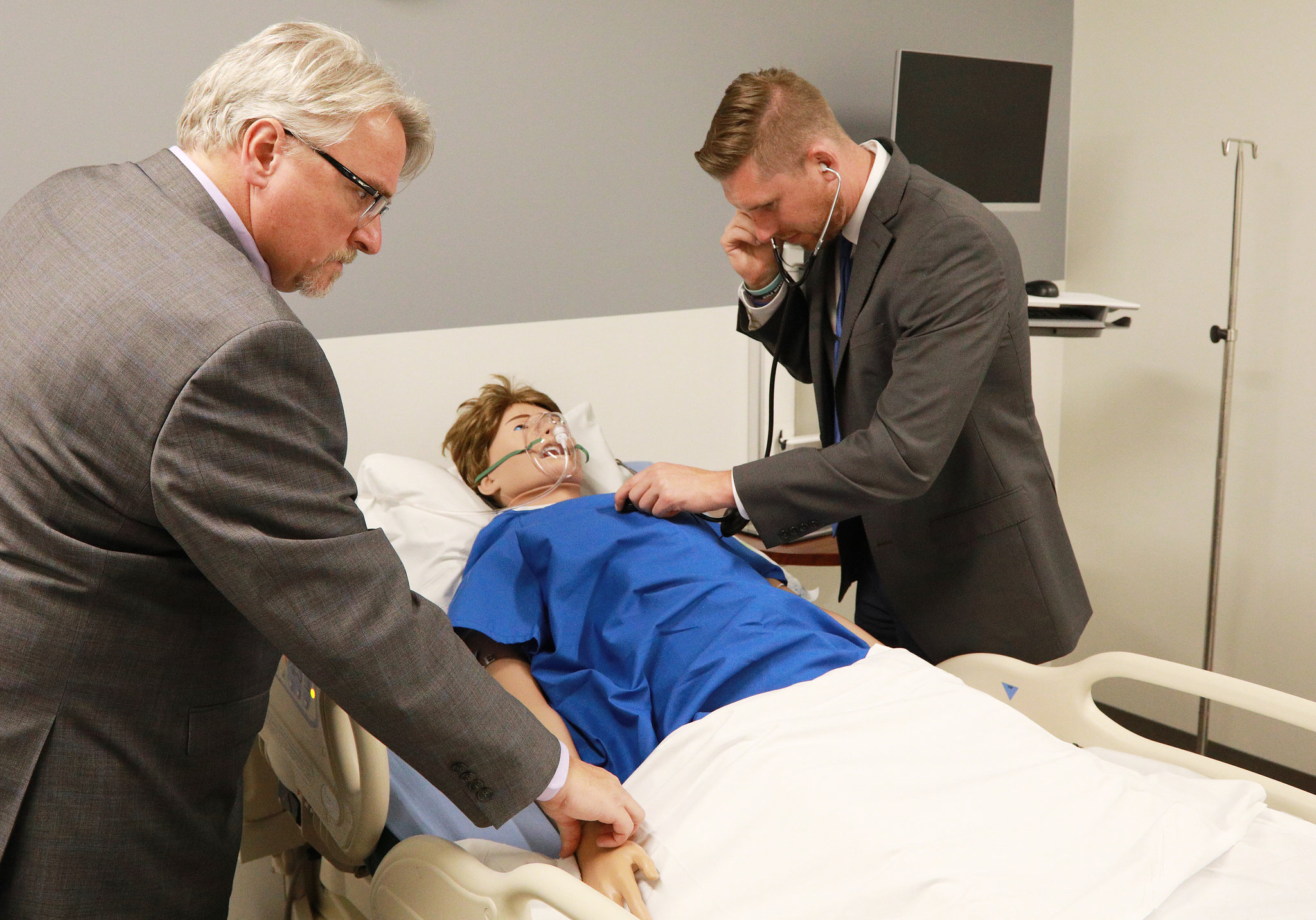 Two men in suits practicing care on a mannequin in a hospital bed