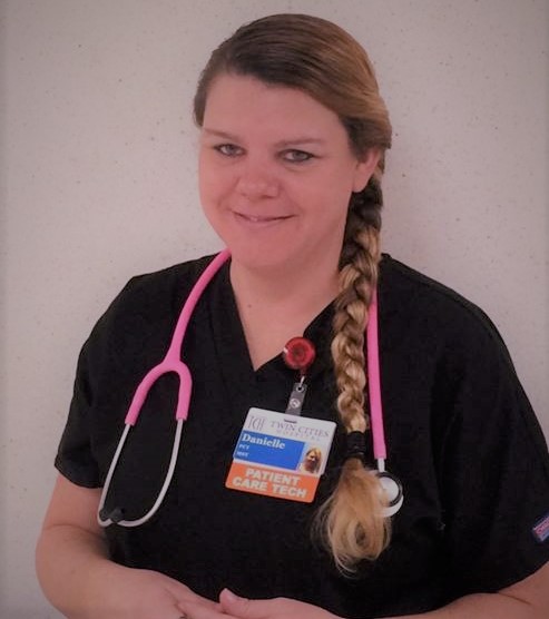A female patient care tech wearing black scrubs with a pink stethoscope around her neck