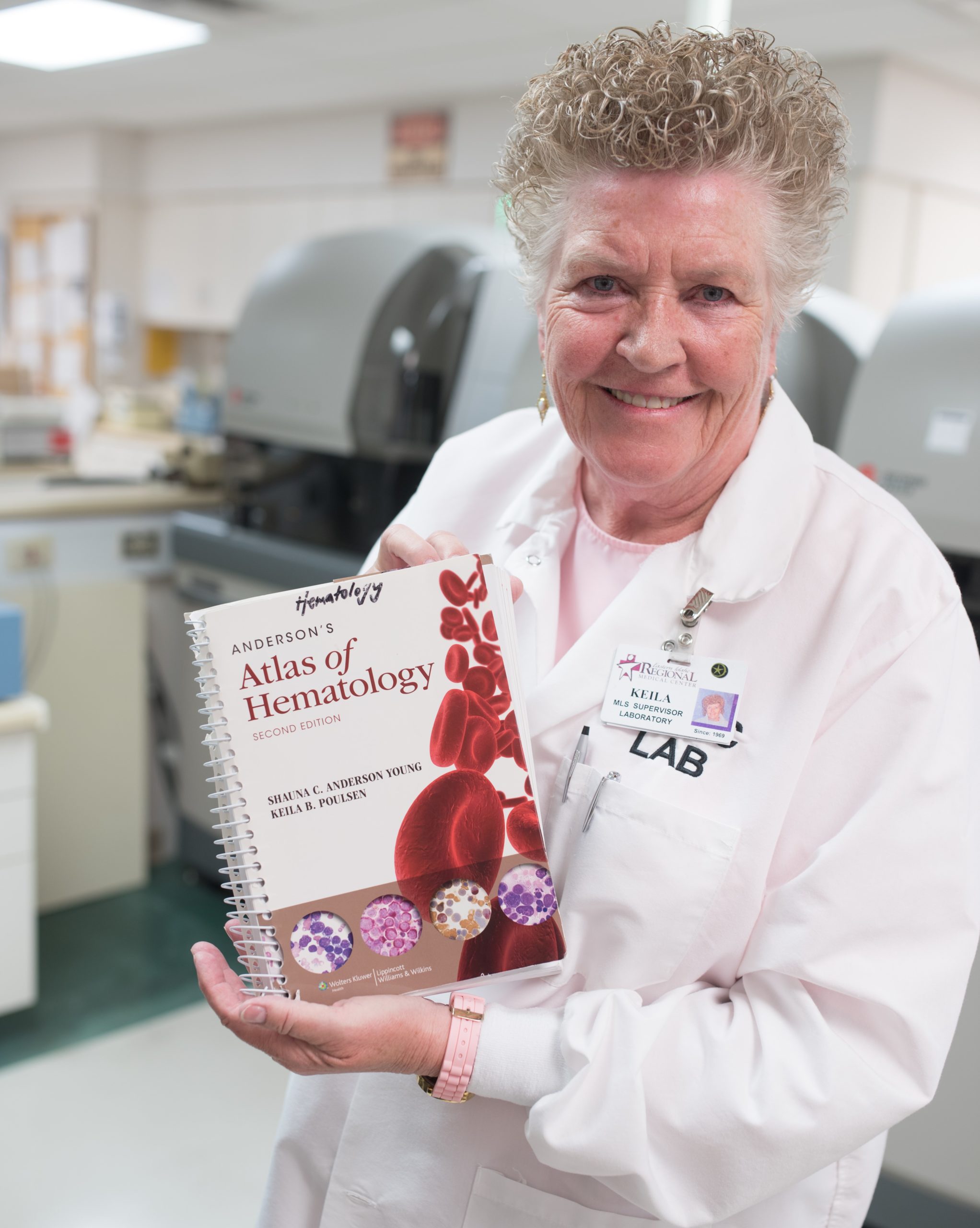 Keila Poulsen standing in a lab, wearing a white lab coat and holding up a book called "Anderson's Atlas of Hematology Second Edition" 