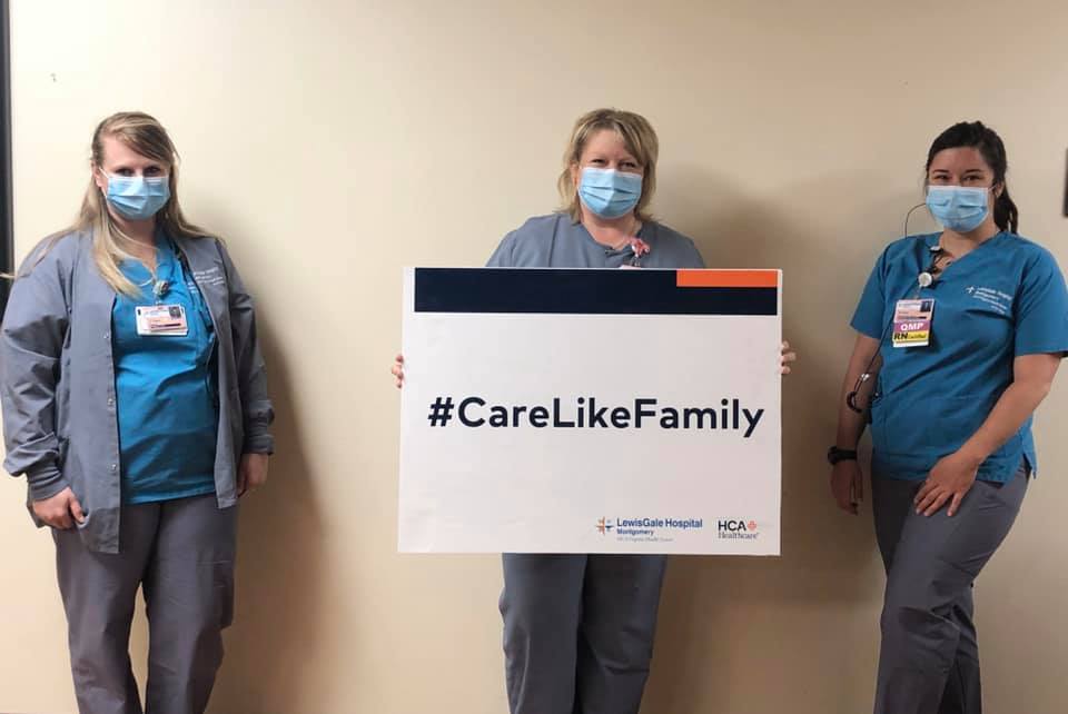 Three female medical professionals with protective face masks and scrubs on, standing in front of a wall holding a sign that says "#CareLikeFamily"