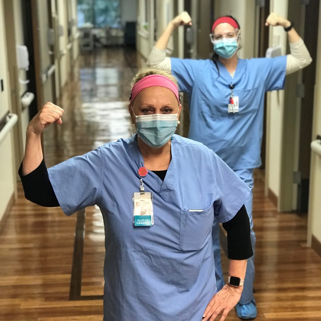 Two female nurses wearing scrubs and protective face masks while flexing their arm muscles in a hallway