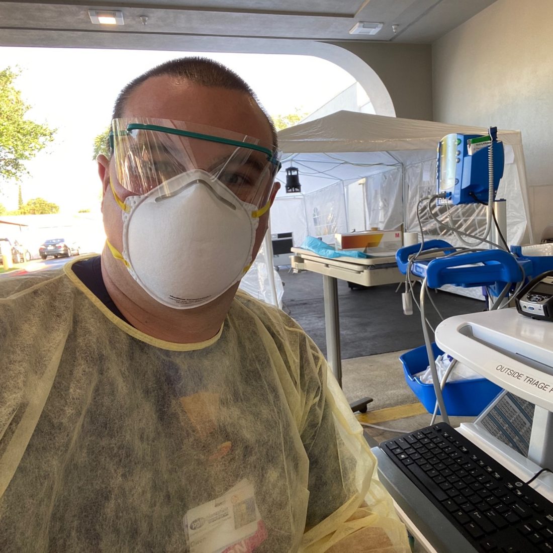 Nurse Owen Rogers wearing protective goggles, mask, and clothing cover over his scrubs, sitting next to medical equipment