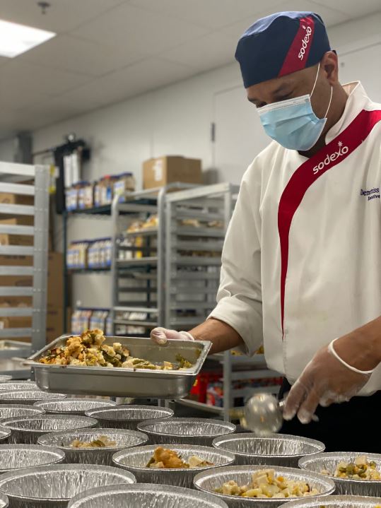 Male chef wearing Sodexo uniform and protective face mask distributes food into aluminum lunch trays