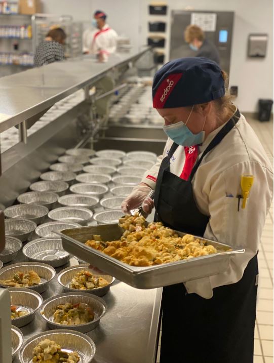 Female chef wearing Sodexo uniform and protective face mask distributes food into aluminum lunch trays