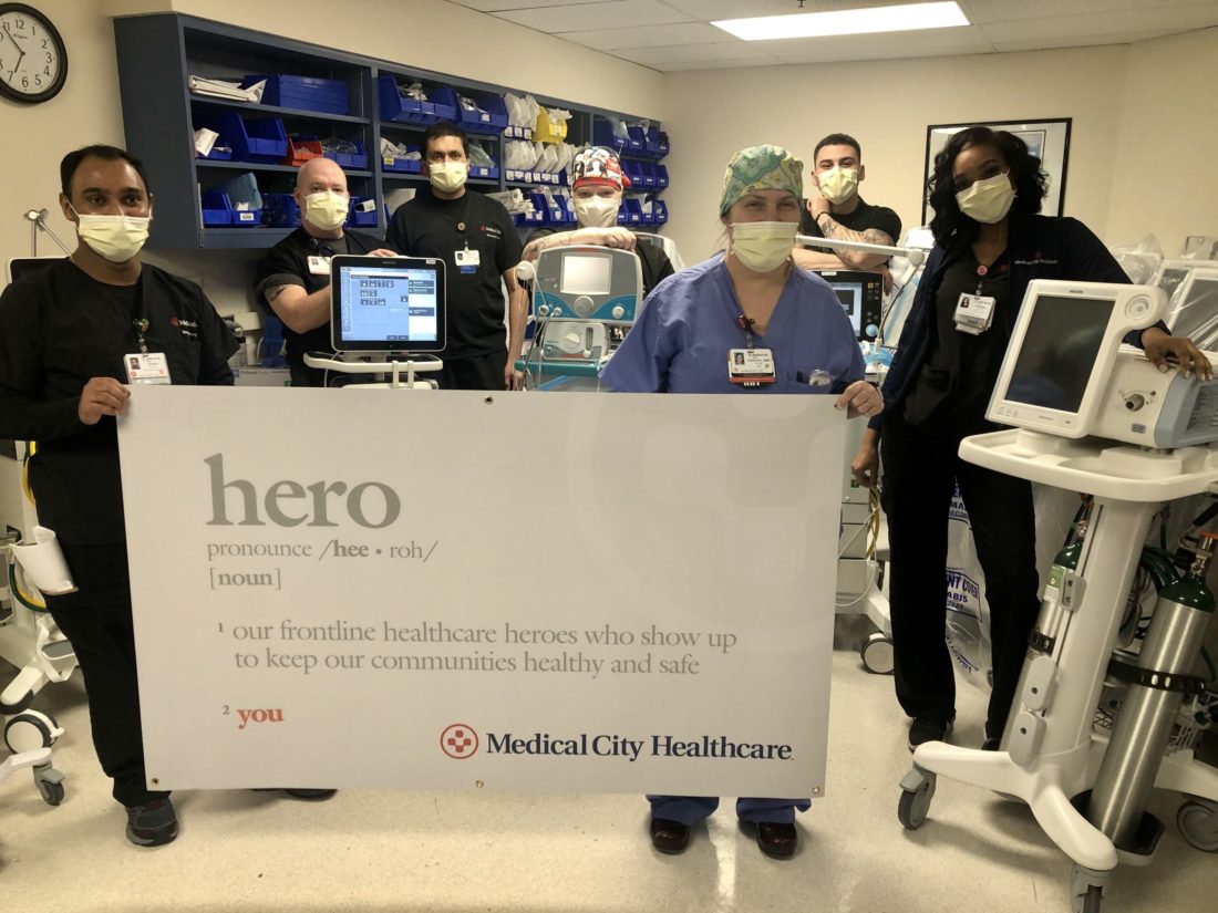 Seven medical professionals wearing scrubs and protective face masks indoors surrounded by medical equipment, and holding up a sign that says "hero pronounce /hee-ro/ [noun] 1 our frontline healthcare heroes who show up to keep our communities healthy and safe 2 you. Medical City Healthcare.
