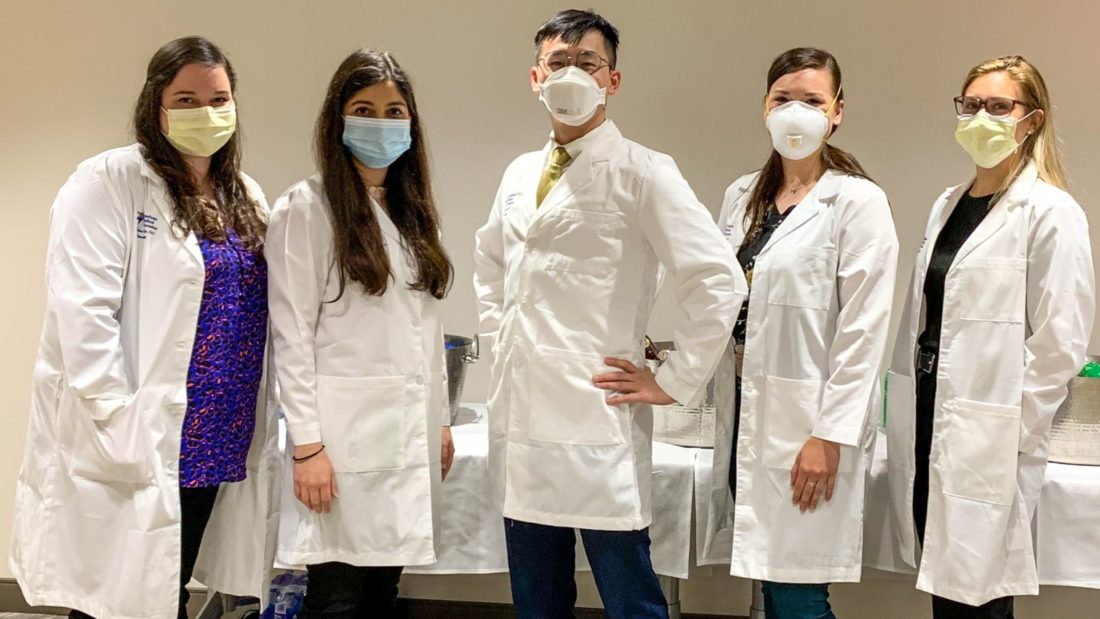 Four women and one man standing together with business clothes, lab coats and protective face masks on