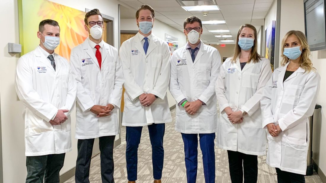 Four men and two women standing together in a hallway, wearing business clothes with lab coats and protective face masks