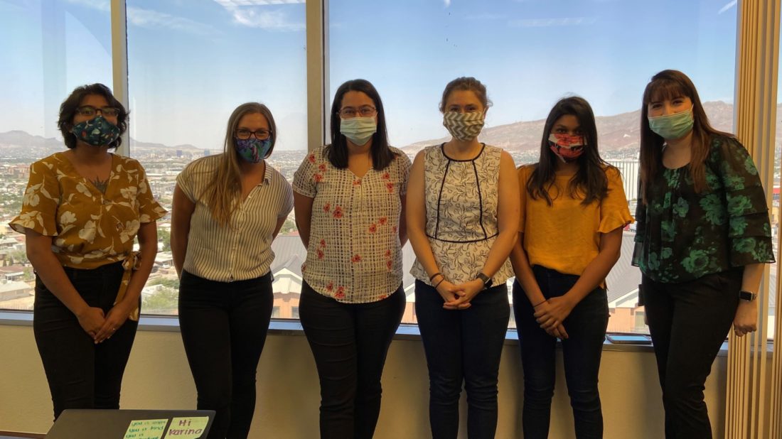 Six women standing in front of a window wearing business casual clothes and protective face masks