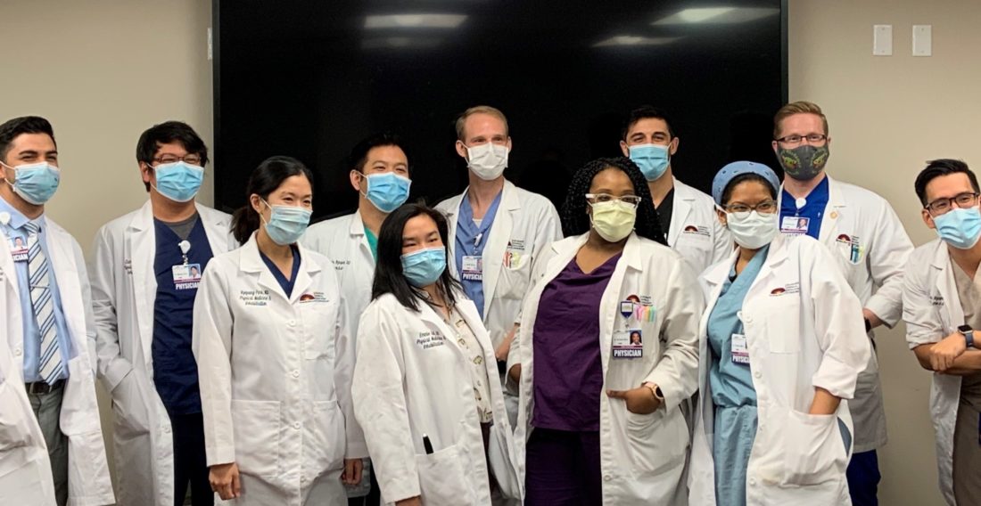 11 adults standing together indoors wearing scrubs, lab coats and protective face masks