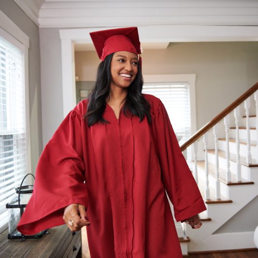 Woman in graduation cap and gown smiling and walking inside home