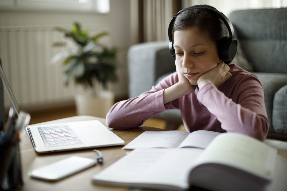 Girl wearing headphones while doing homework with laptop, textbook and notebook 