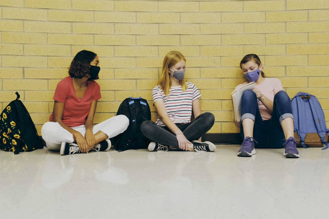Three female high school students wearing face masks sitting on the ground with their backpacks. 
