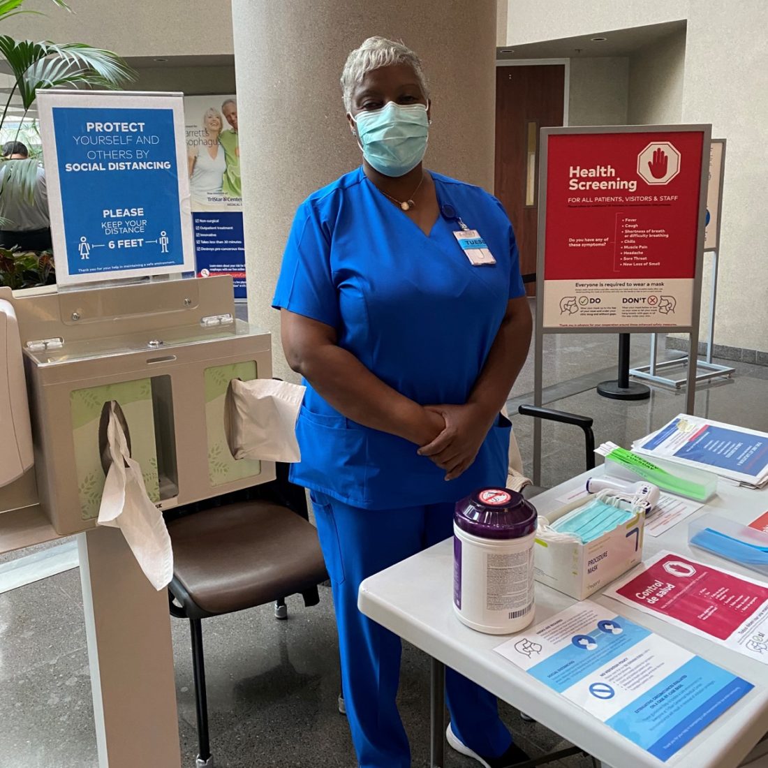 Woman wearing face mask and scrubs behind table with health screening materials.