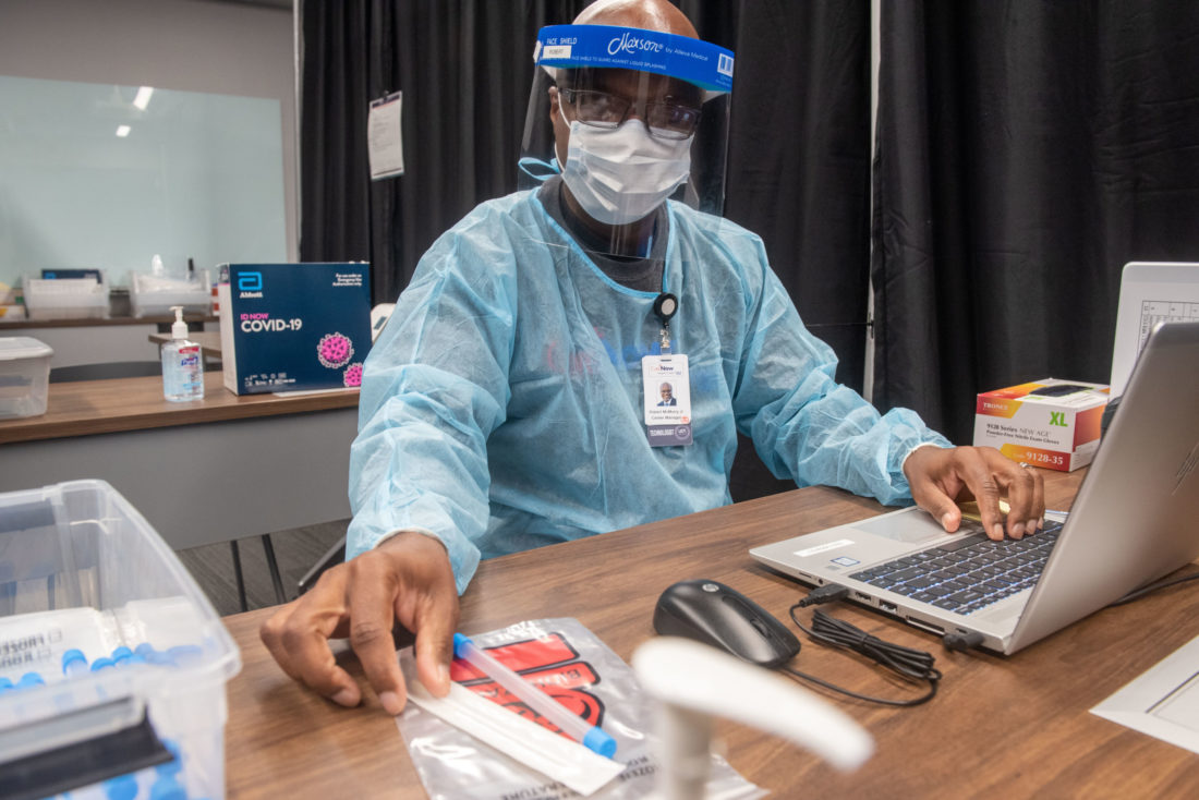 Man wearing personal protective equipment sitting at a desk with a laptop and testing materials. 