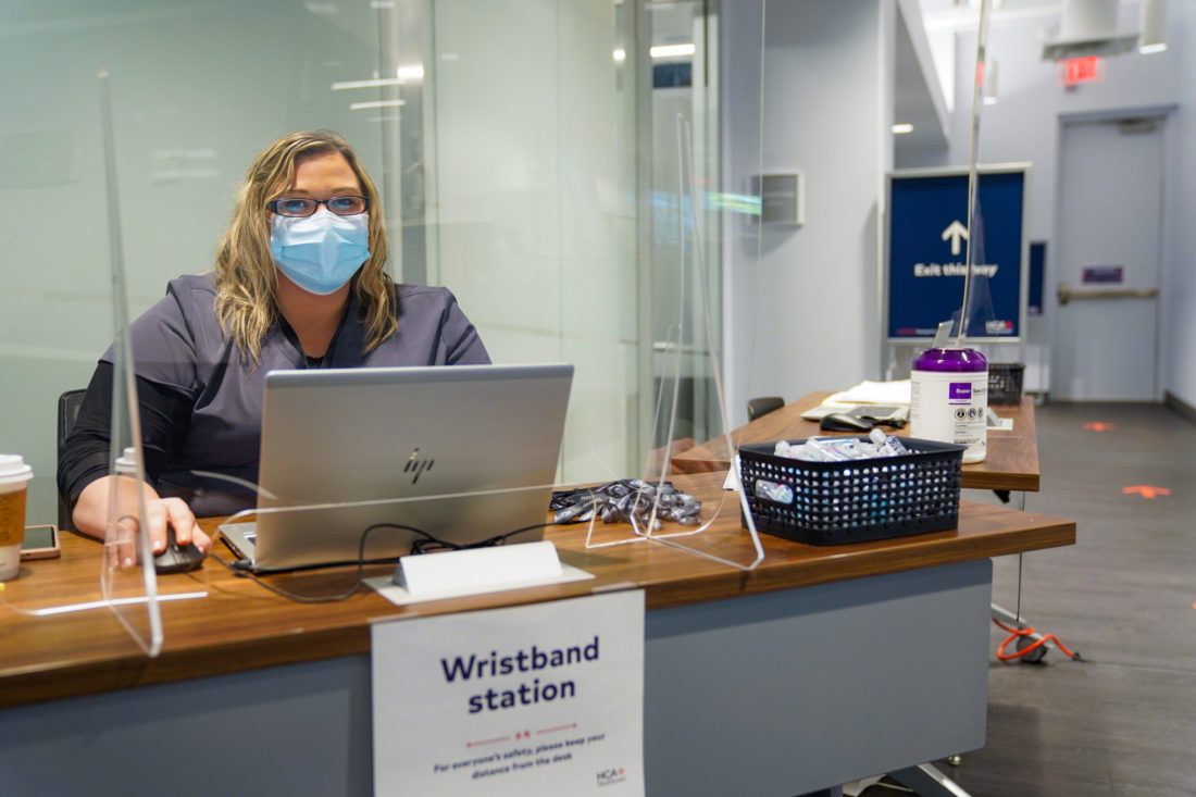 Woman wearing face mask and scrubs sitting at a desk with a laptop behind a clear plastic shield. 