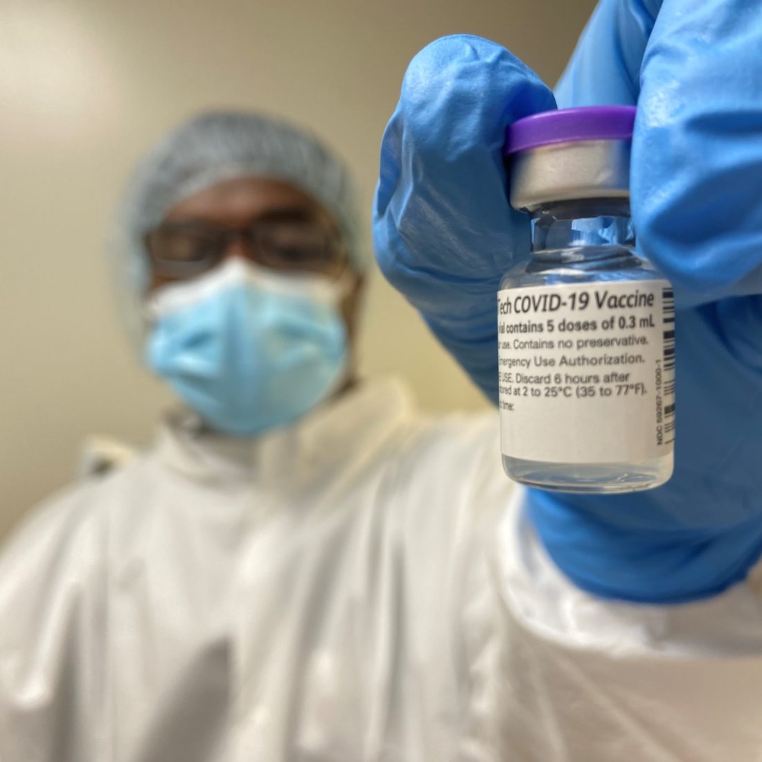 Pharmacy technician wearing personal protective equipment holding a vial of the COVID-19 vaccine