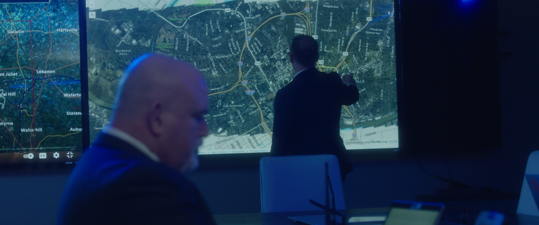 A man wearing a suit looks at a map on a large screen while another man works on a computer nearby