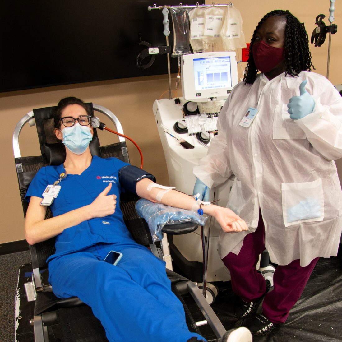 Two female healthcare workers giving thumbs up sign as one donates blood
