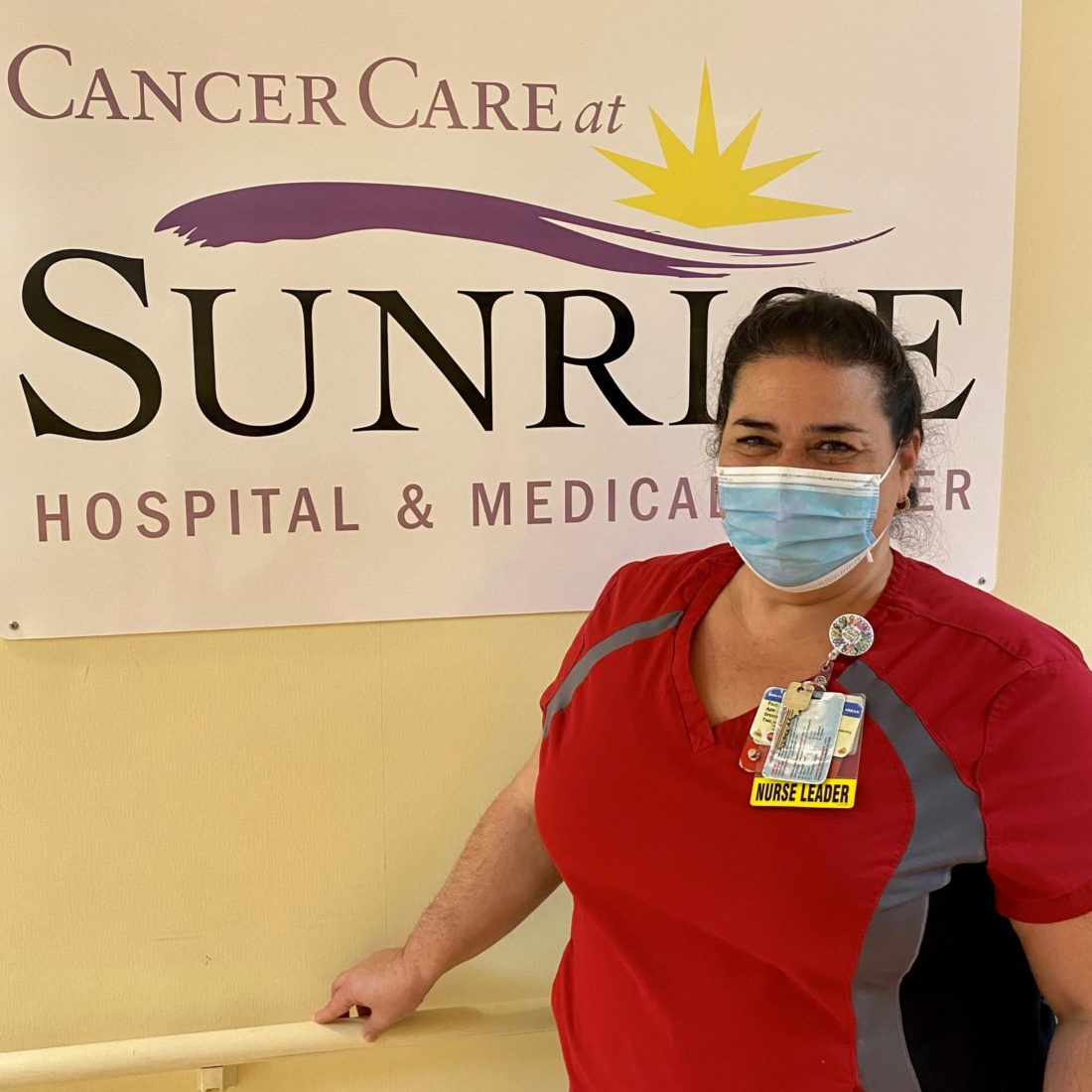 A female nurse wearing scrubs and a face mask standing in front of hospital sign. 