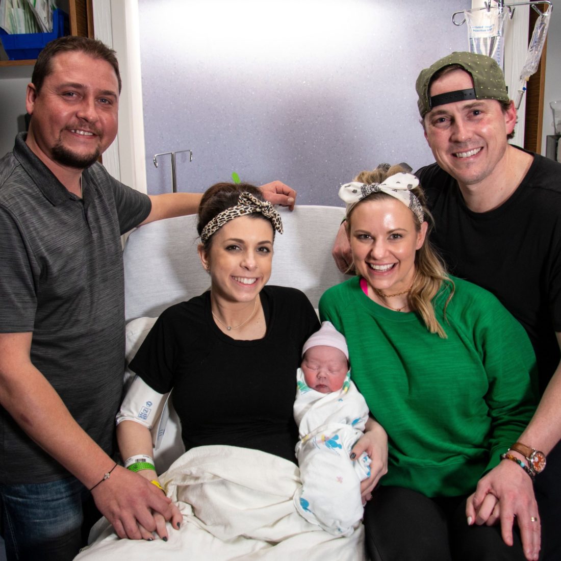 Woman in hospital bed holding newborn baby with another woman and two men surrounding her. 
