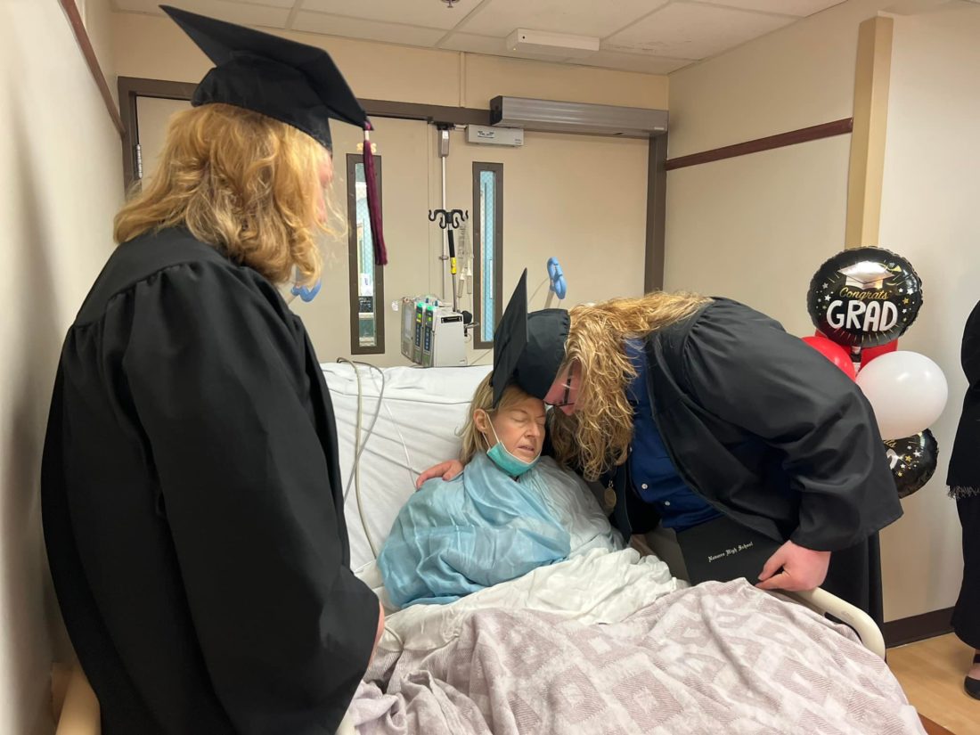 Two teenagers in graduation caps and gowns hugging a woman in a hospital bed