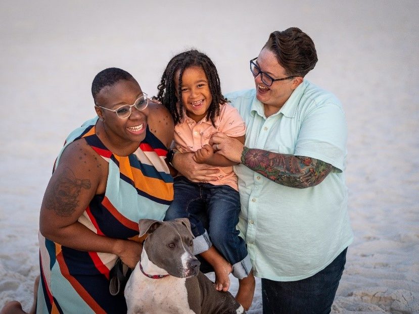 Kadesha, spouse Meg and son laughing on the beach with their dog.