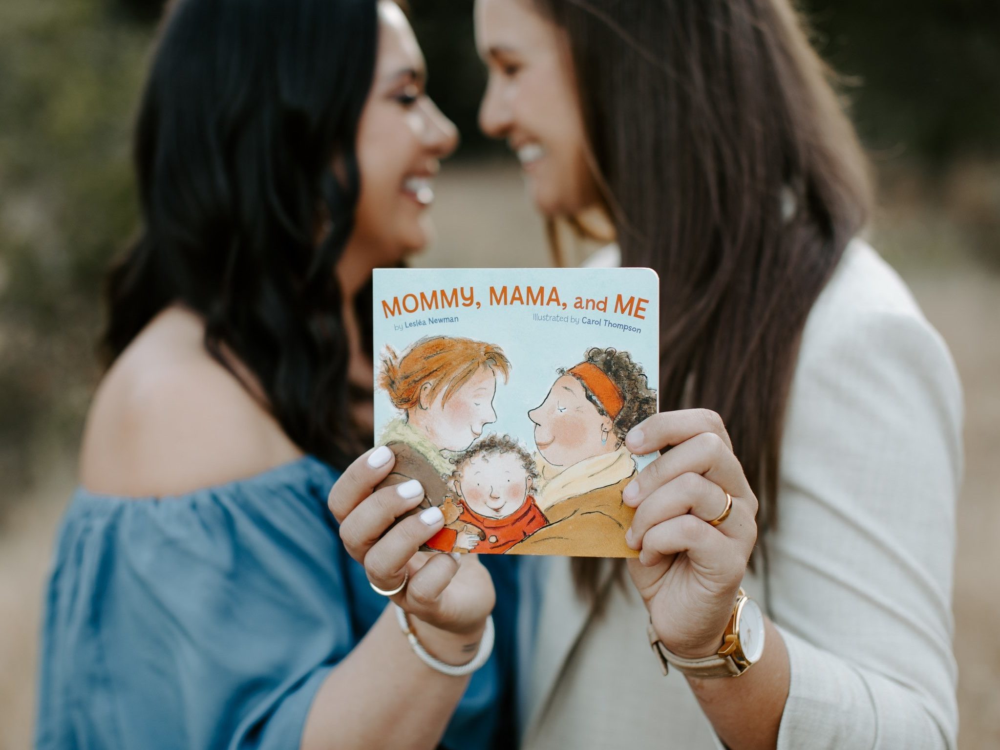 Jaime and her wife, Jessica, holding up a book that reads "Mommy, Mama, and Me." 