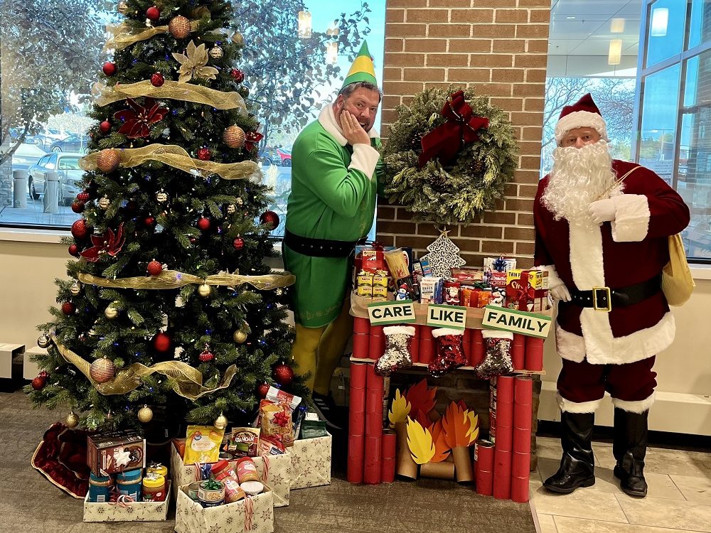 Santa and Buddy the Elf standing around a fireplace made of donated canned goods. 